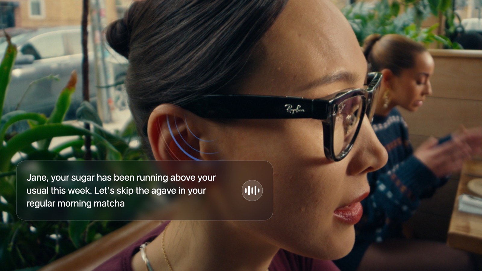 Close-up of a woman wearing Ray-Ban Meta glasses in a café, a soundwave icon by her ear showing Oremi speaking: “Jane, your sugar has been running above your usual this week. Let's skip the agave in your regular morning matcha.”