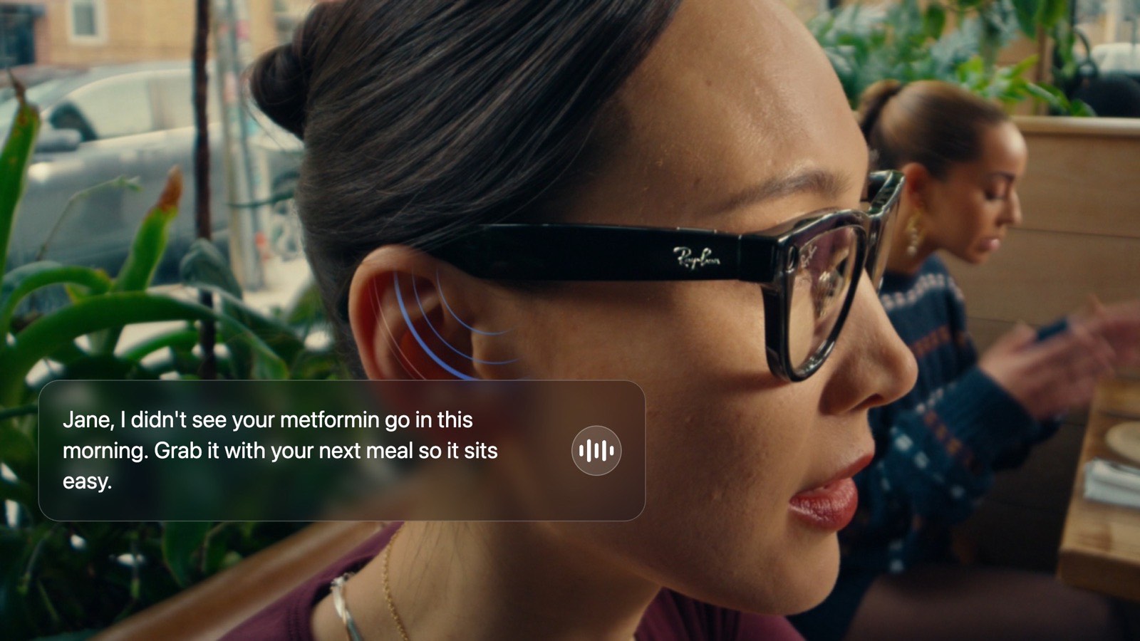 Close-up of a woman wearing Ray-Ban Meta glasses in a café, a soundwave icon by her ear showing Oremi speaking: “Jane, I didn't see your metformin go in this morning. Grab it with your next meal so it sits easy.”