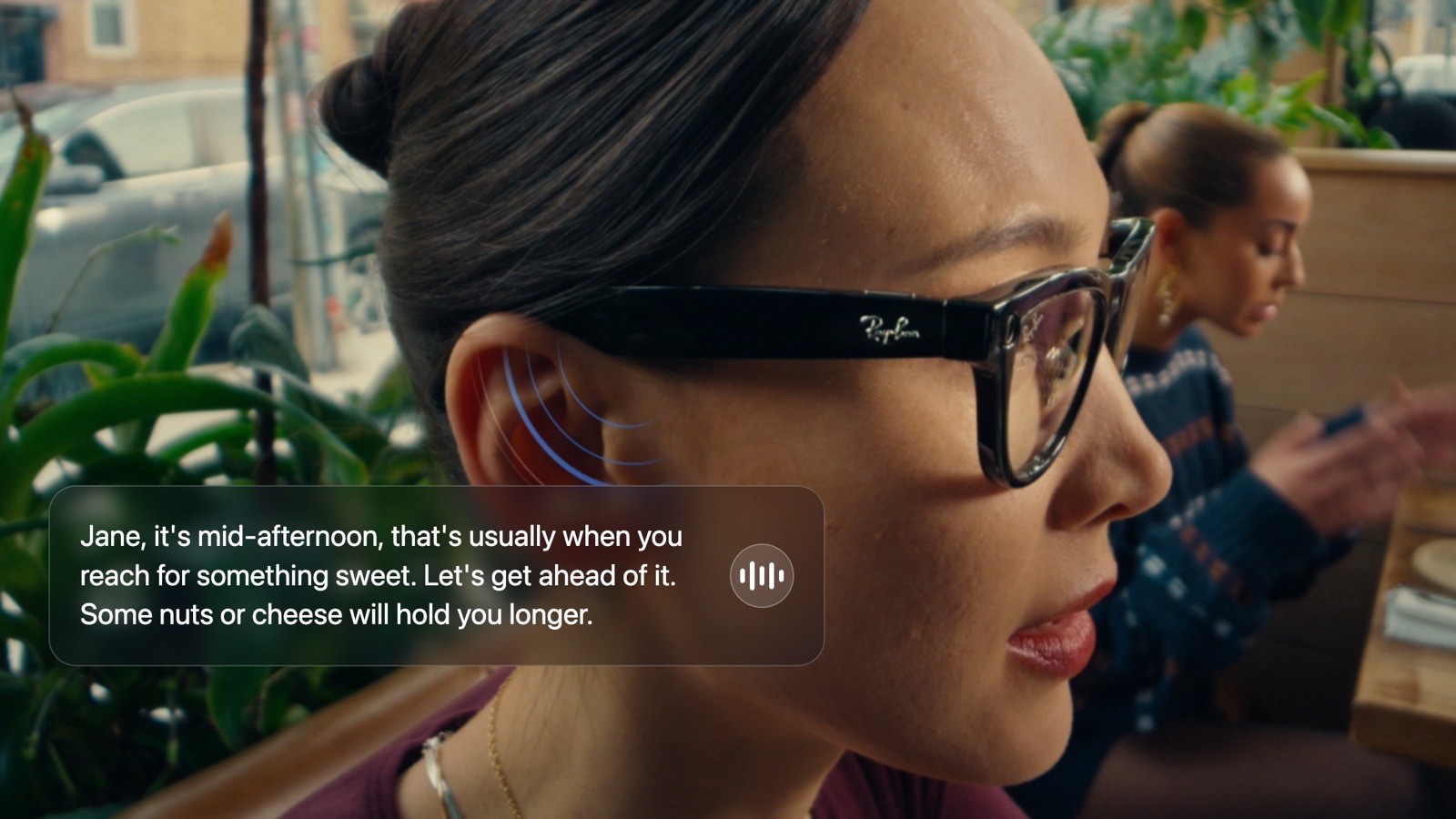 Close-up of a woman wearing Ray-Ban Meta glasses in a café, a soundwave icon by her ear showing Oremi speaking: “Jane, it's mid-afternoon, that's usually when you reach for something sweet. Let's get ahead of it. Some nuts or cheese will hold you longer.”